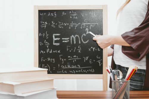 Photo of teacher's hand pointing to chalkboard with E=mc squared written in the center.