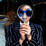 Photo of a person dressed in a wide white pinstripe on navy suit and wearing big 80s type sunglasses, looking through a magnifying glass.