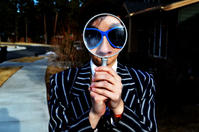 Photo of a person dressed in a wide white pinstripe on navy suit and wearing big 80s type sunglasses, looking through a magnifying glass.