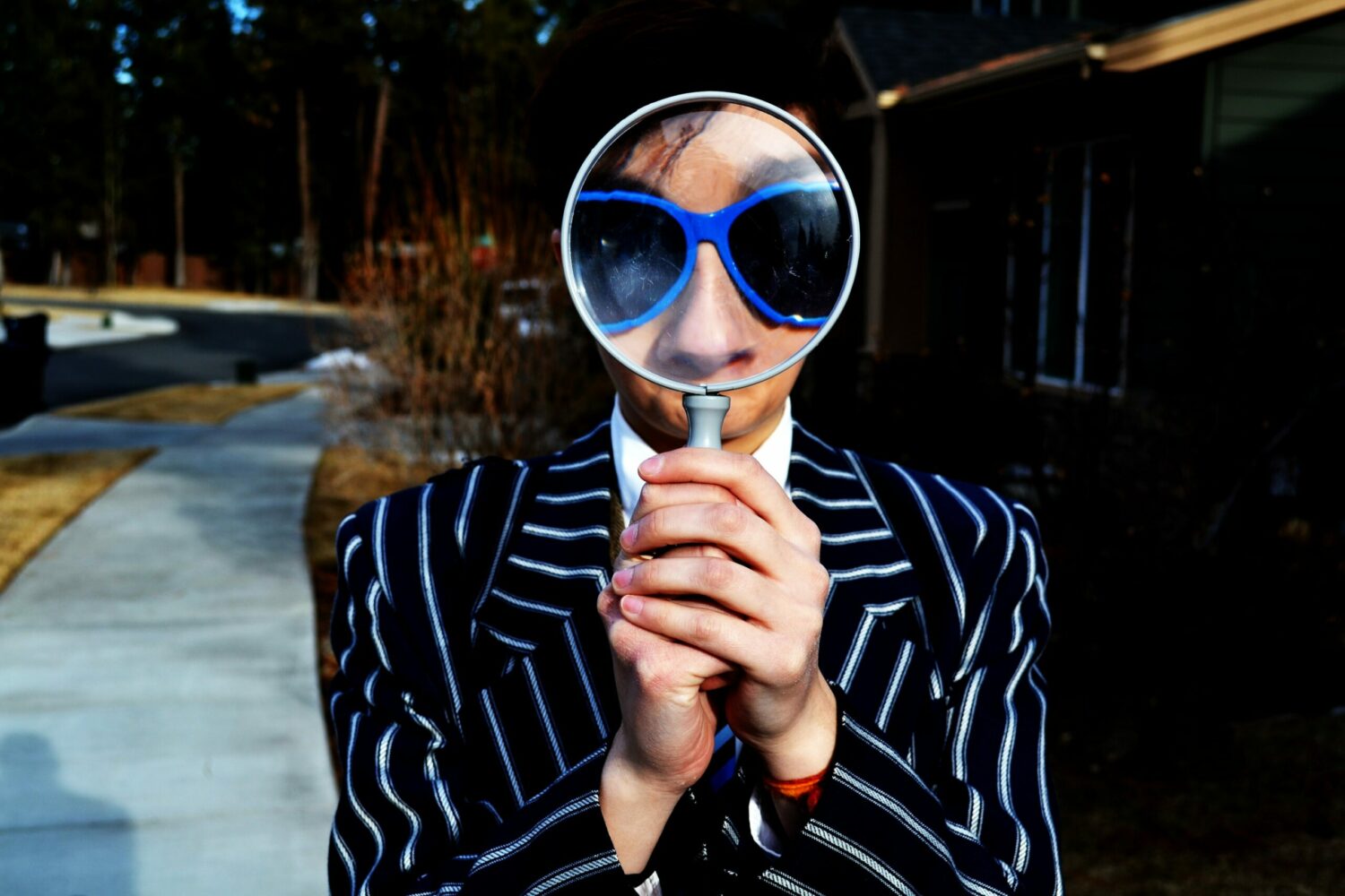 Photo of a person dressed in a wide white pinstripe on navy suit and wearing big 80s type sunglasses, looking through a magnifying glass.